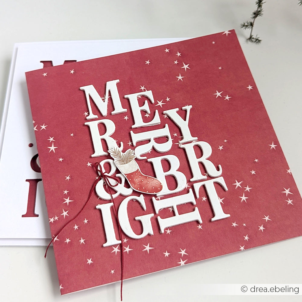Rote Weihnachtskarte, gestaltet mit Alexandra Renke Designpapier Sternenzauber Bordeauxrot, mit "MERRY & BRIGHT" in Weiß, Sternen und einem Strumpfschmuck mit Schnur - ideal für Papierprojekte oder Buchbinderei.