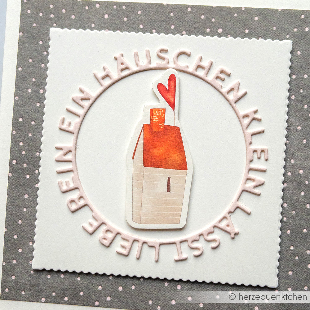Ein kleines Häuschen mit rotem Herzschornstein und deutschem Text auf gepunktetem, quadratischem Hintergrund - ideales Alexandra Renke Designpapier für Buchbinderei oder ausgefallene Papierprojekte.