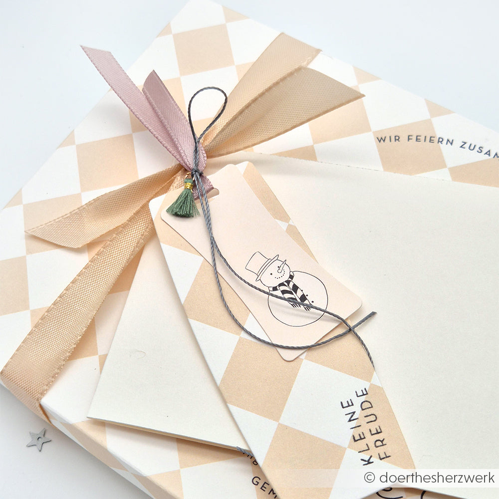 Verpackt in Alexandra Renke's Designpapier Großes Karo beige mit cremefarbenem Rautenmuster und beigem Band, veredelt mit einem Schneemann-Anhänger - eine charmante Papierprojekte-Idee. Der Umschlag aus der Buchbinderei Tradition liegt darunter.
