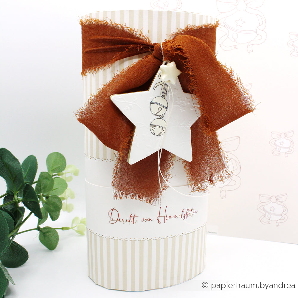 Ein hoher gestreifter Geschenkkarton von Alexandra Renke aus hochwertigem Designpapier, verziert mit einer braunen Stoffschleife und einem weißen Sternenornament, steht neben einer grünen Blattpflanze.