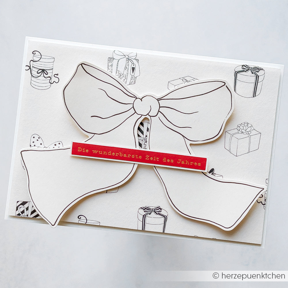 Eine Karte von Alexandra Renke mit einer großen konturierten Schleife und deutschem Text, umgeben von Geschenkskizzen - ideal für Papierprojekte oder um Ihrer nächsten buchbinderischen Kreation ein besonderes Flair zu verleihen.