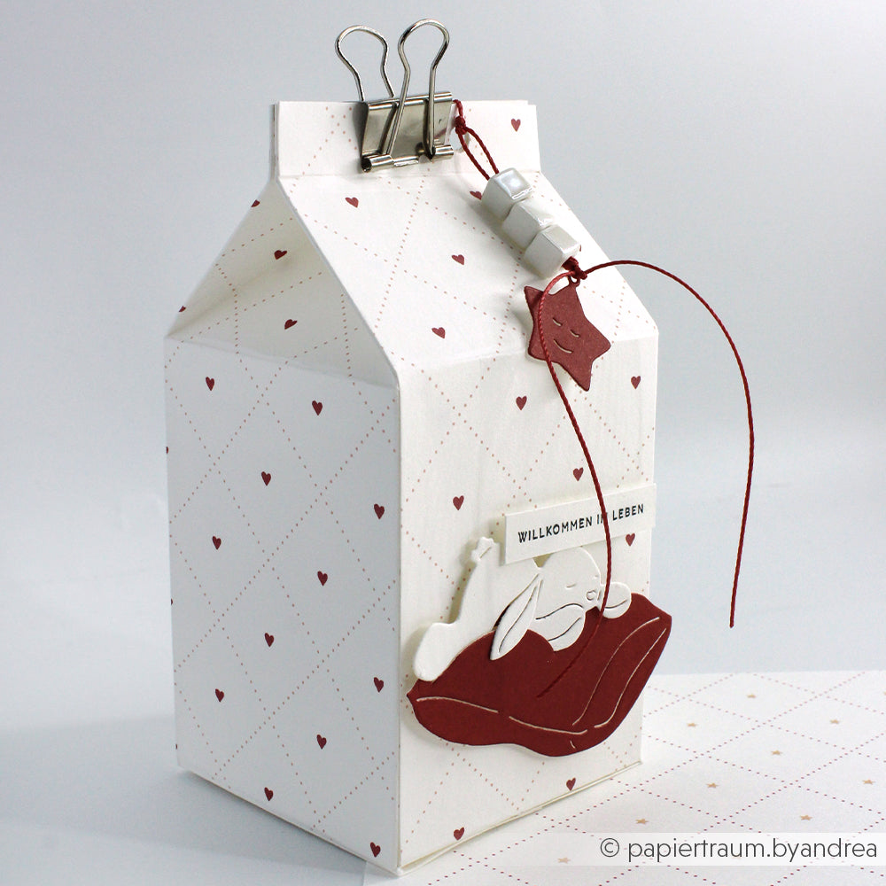 Die weiße Milchtüte aus Papier von Alexandra Renke hat ein rotes Herzmuster, das sich ideal für Papierprojekte eignet. Sie ist mit Anhängern und Akzenten aus Designpapier verziert und wird von einer Heftklammer gekrönt.