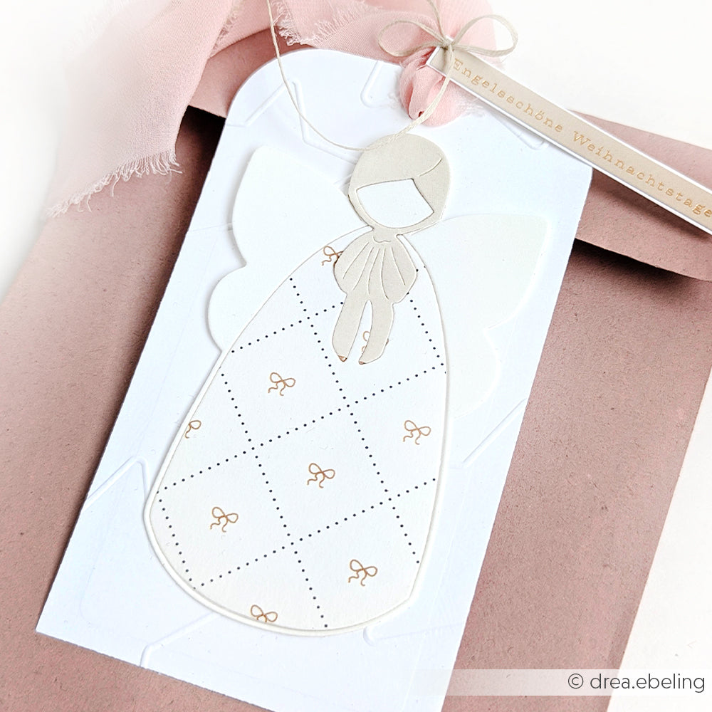 Minimalistischer Engel-Geschenkanhänger von Alexandra Renke auf Designpapier, präsentiert auf einem rosa Umschlag mit Schleife und Etikett. Ideal für Papierprojekte oder Buchbinderei-Fans, die stilvolle Details suchen.