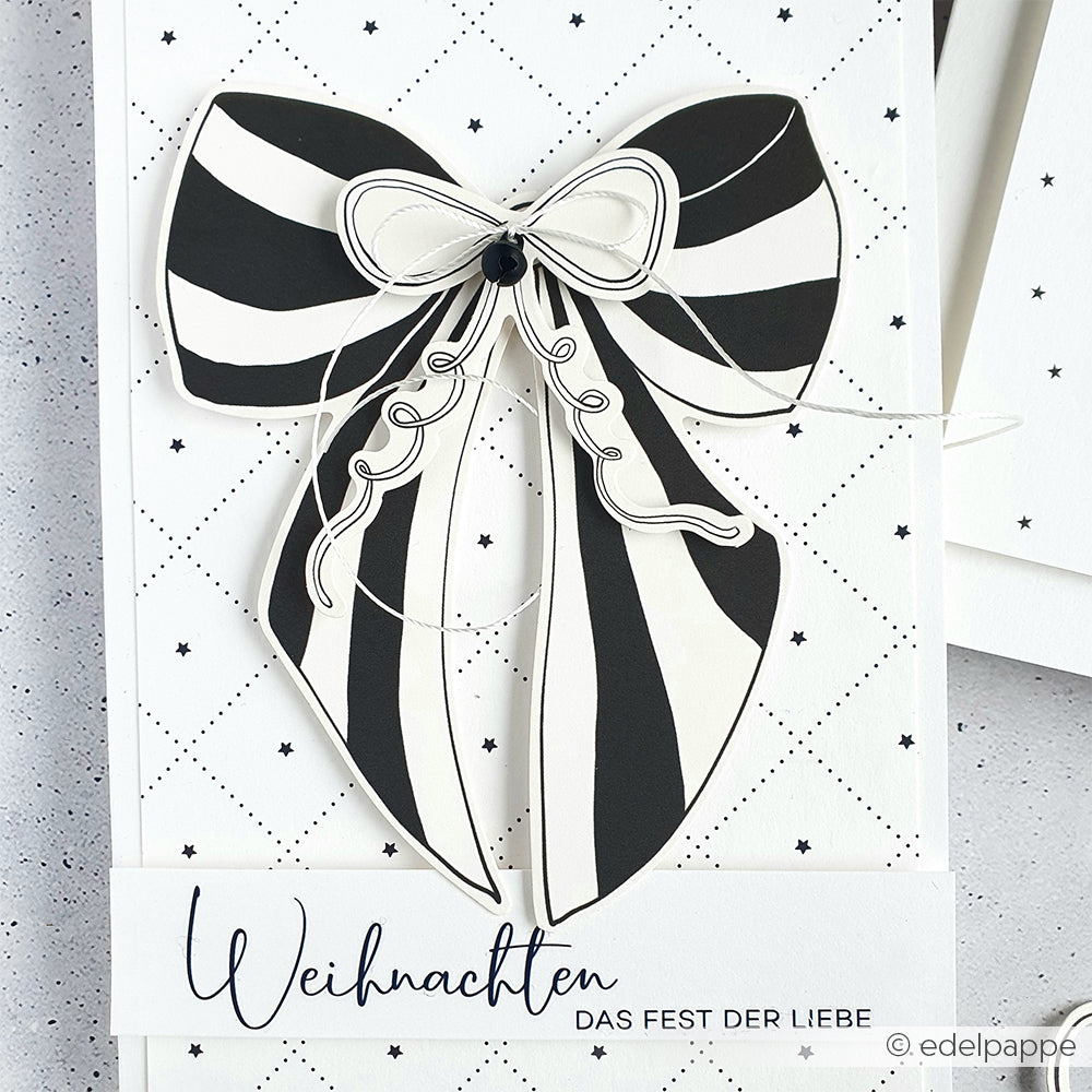 Schwarz-weiß gestreifte Schleife auf einer festlichen Karte mit Alexandra Renke Designpapier, mit dem Text "Weihnachten Das Fest der Liebe" am unteren Rand.