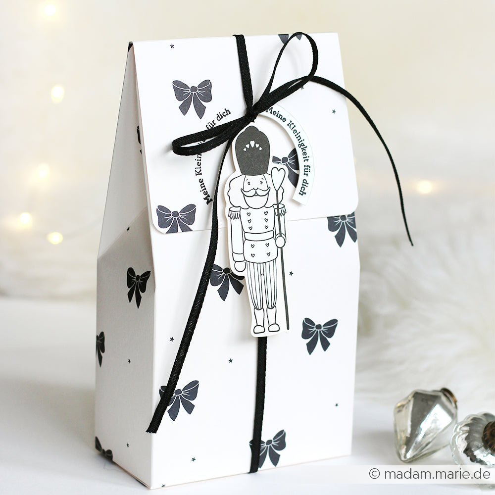 Weiße Geschenktüte von Alexandra Renke mit schwarzem Schleifenmuster, verschnürt mit schwarzer Kordel und Nussknacker-Anhänger - ideal für Papertie-Projekte oder um Ihre Weihnachtsgeschenke mit charmanten Ephemera zu ergänzen.