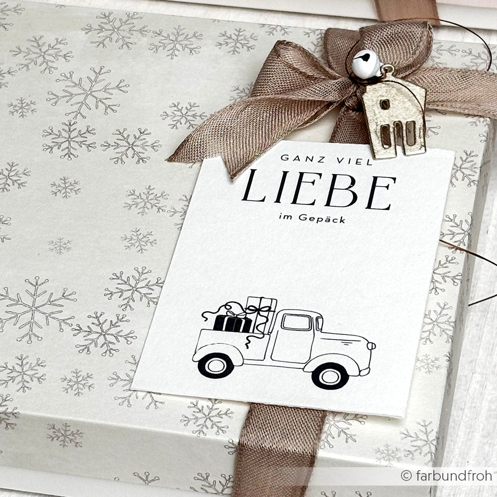 Geschenkverpackung aus Alexandra Renke-Schneeflockenpapier mit einer Karte, auf der ein Lastwagen mit Geschenken und ein deutscher Text aus "Liebe im Gepäck" abgebildet ist, verziert mit Schleife und Anhängern - perfekt für festliche Papierprojekte.