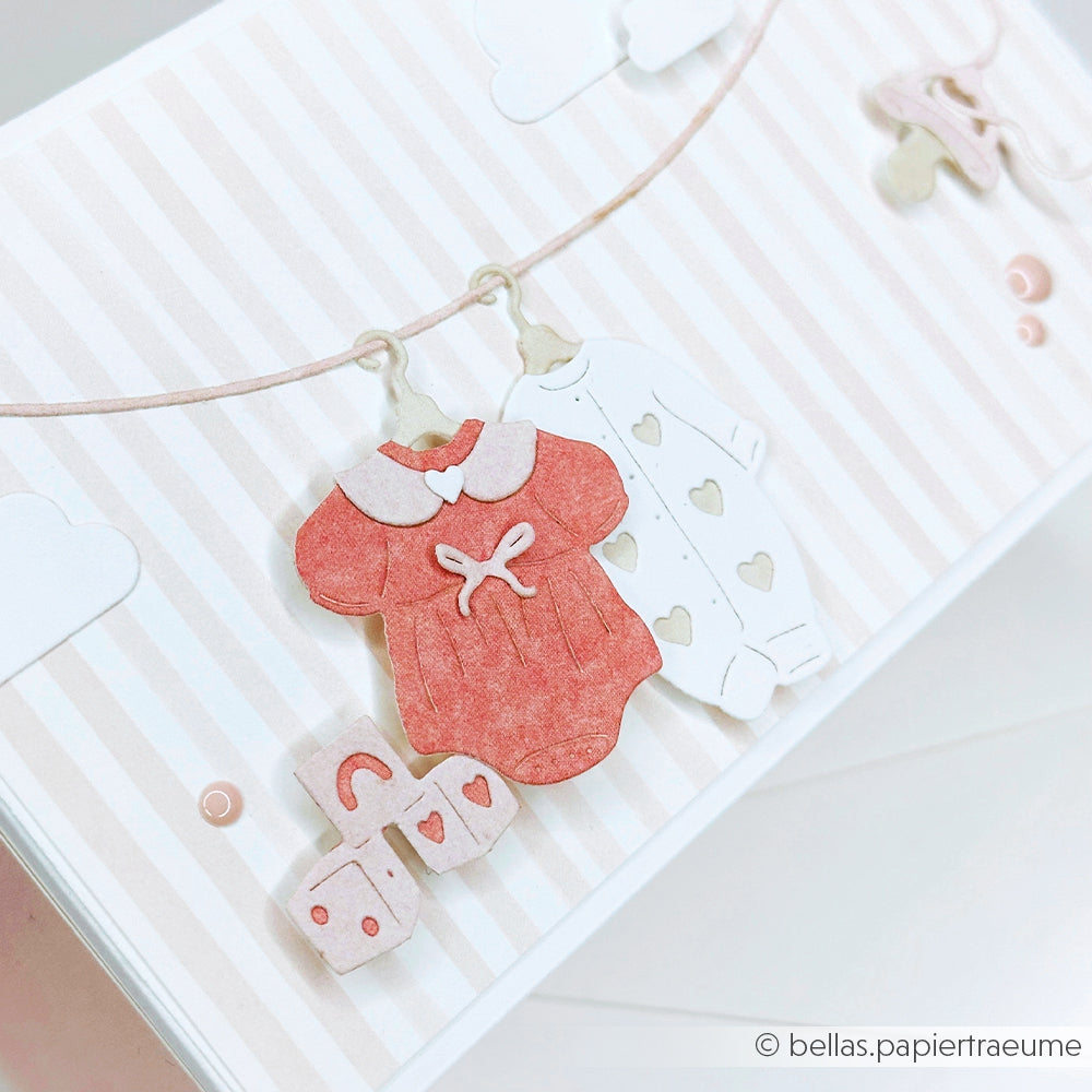 Auf einer Leine vor einem pastellrosa gestreiften Hintergrund hängen Papierausschnitte von Babykleidung und Bauklötzen, hergestellt mit dem charmanten Designpapier von Alexandra Renke.