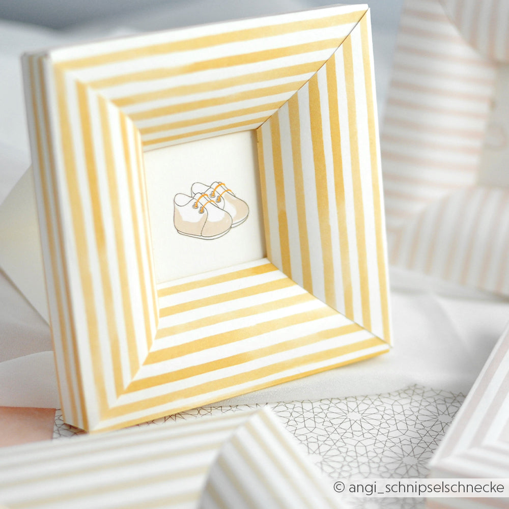 Dekorativer Bilderrahmen von Alexandra Renke aus Designpapier Streifen senfgelb mit einer Illustration von Babyschuhen - ideal für Papierprojekte oder Buchbinderei auf hellem Hintergrund.
