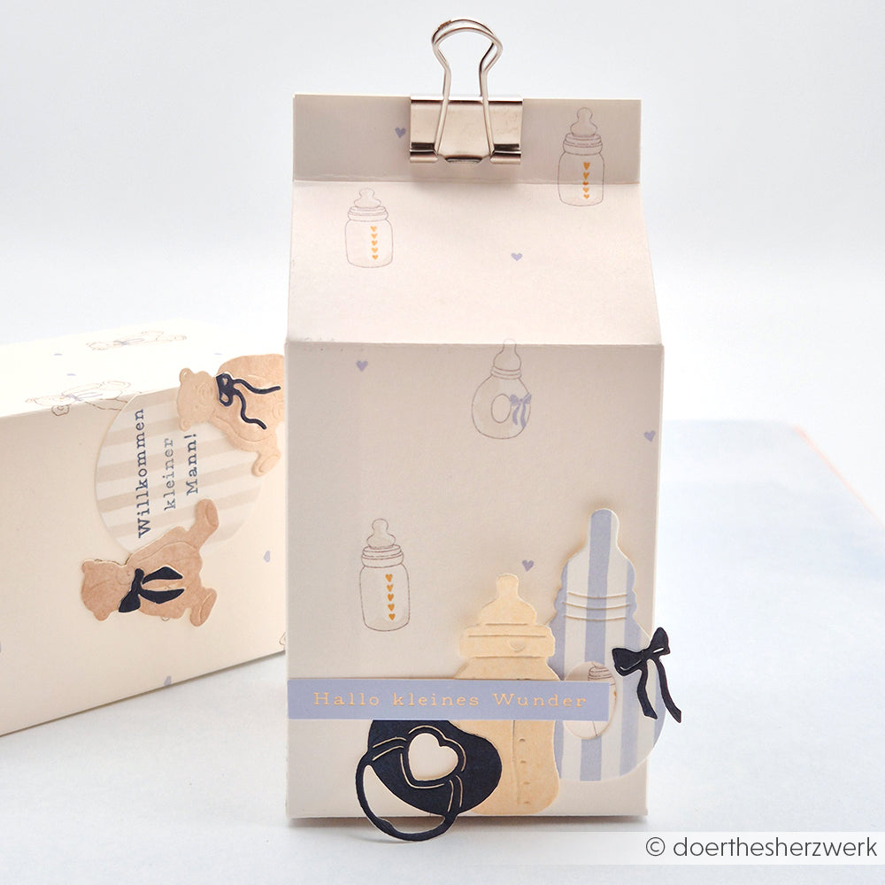Die cremefarbene Baby-Geschenkbox von Alexandra Renke ist mit Designpapier gestaltet, mit Flaschen- und Schnuller-Motiven versehen und mit einer Papierschleife und einem Clip versehen - perfekt für Papierprojekte oder Buchbinderei-Geschenke.