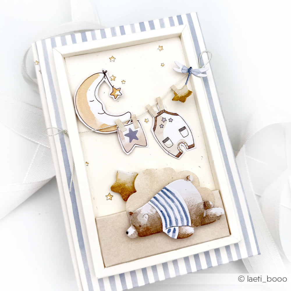 Ein niedlicher Bär im Schlafanzug schläft unter Sternen, einem Mond und einem kleinen Hausdekor in einem gestreiften Rahmenkarton - perfekt für Papierprojekte mit Alexandra Renke Designpapier Sternenhimmel-Akzenten.