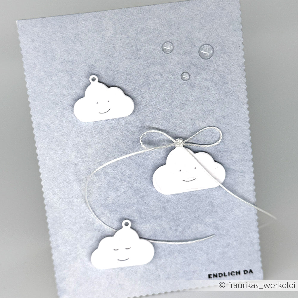Drei weiße Wolkenanhänger mit Gesichtern und einer silbernen Schleife ruhen auf einer grauen, strukturierten Alexandra Renke Designpapier-Karte, perfekt für Papierprojekte, mit ENDLICH DA am Boden.
