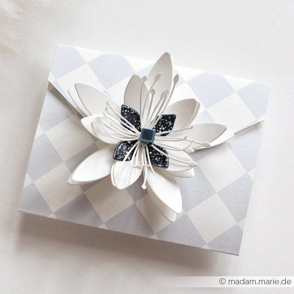 Ein gefalteter Umschlag von Alexandra Renke aus Designpapier zeigt eine weiße und schwarze Papierblume auf einem grauen Hintergrund mit Rautenmuster.