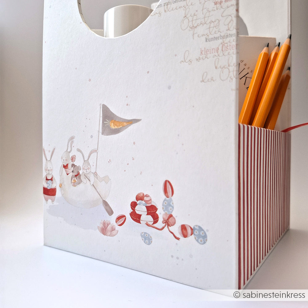 Weiße Schachtel mit roten Streifen von Alexandra Renke, verziert mit Hasen und Eiern und gefüllt mit Bleistiften - ideal für Papierprojekte oder als Geschenkbox für Grußkarten. Eine festliche Ergänzung zu Ihrer Designpapier Ostern Kollektion.