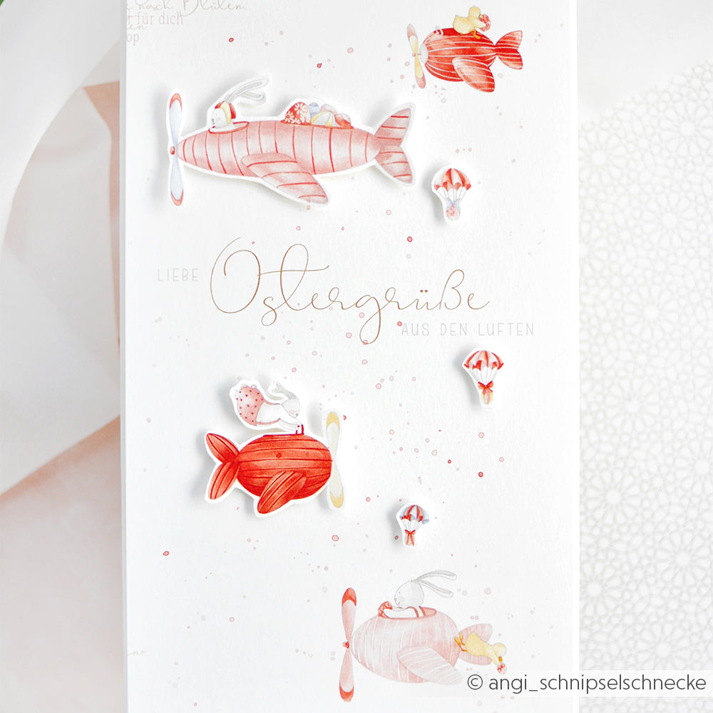 Oster-Grußkarte von Alexandra Renke mit roten Flugzeugen und Häschen mit Hüten auf Designpapier. Text: Liebe Ostergrüße aus den Lüften. Ideal für besondere Ostergrüße und liebevolle Papierprojekte.