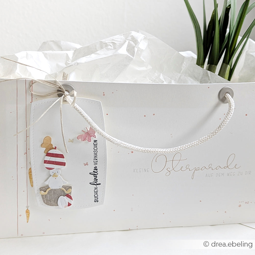 Weiße Geschenktüte von Alexandra Renke mit einem dekorativen Osteranhänger aus Osterparade-Designpapier, Hasenmotiv, Seidenpapier und einer Pflanze im Hintergrund - ideal für kreative Papierprojekte oder Ihr nächstes Buchbinderei-Projekt.