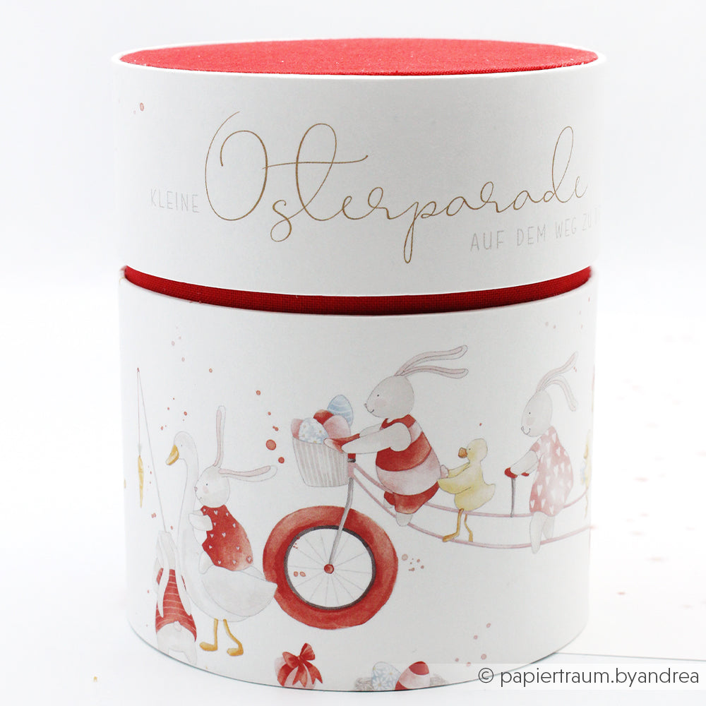 Runder weißer Geschenkkarton mit rotem Deckel von Alexandra Renke mit Hasen und einer Ente in Festtagskleidung auf einem Fahrrad, hergestellt mit Buchbinderei-Technik und skurrilem Osterparade-Designpapier - perfekt für kreative Papierprojekte.