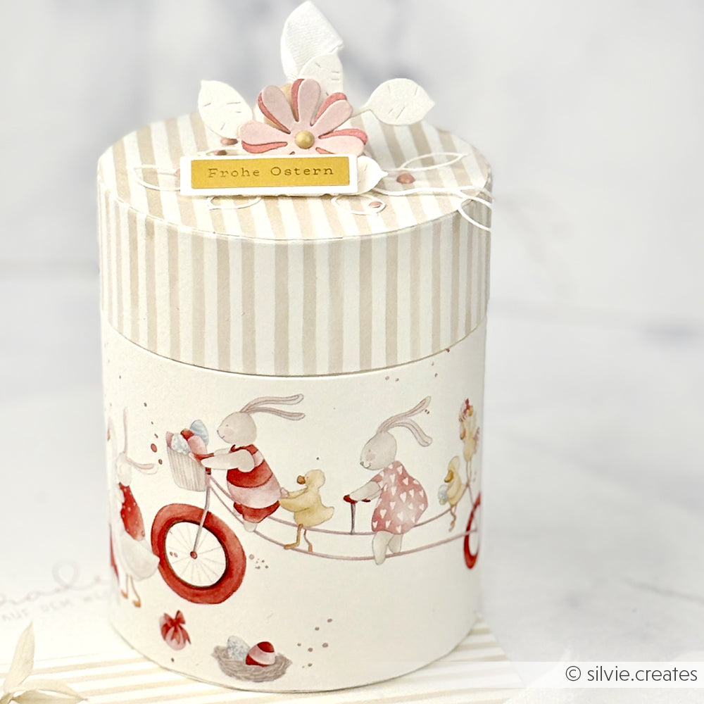 Runde Geschenkschachtel mit gestreiftem Deckel, verziert mit Alexandra Renke Designpapier Osterparade, mit niedlichen Hasen, Enten und einem Frohe Ostern-Anhänger - ideal für Papierprojekte oder als Ergänzung zu Ihren Grußkarten.