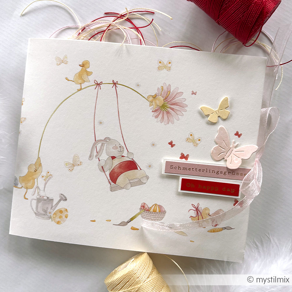 Handgemachte Karte von Alexandra Renke mit einem Hasen auf einer Schaukel, Schmetterlingen, Blumen und Bändern. Gefertigt aus Designpapier Osterglück und verziert mit dem Gruß Schmetterlingsgrüße - perfekt für besondere Papierprojekte.