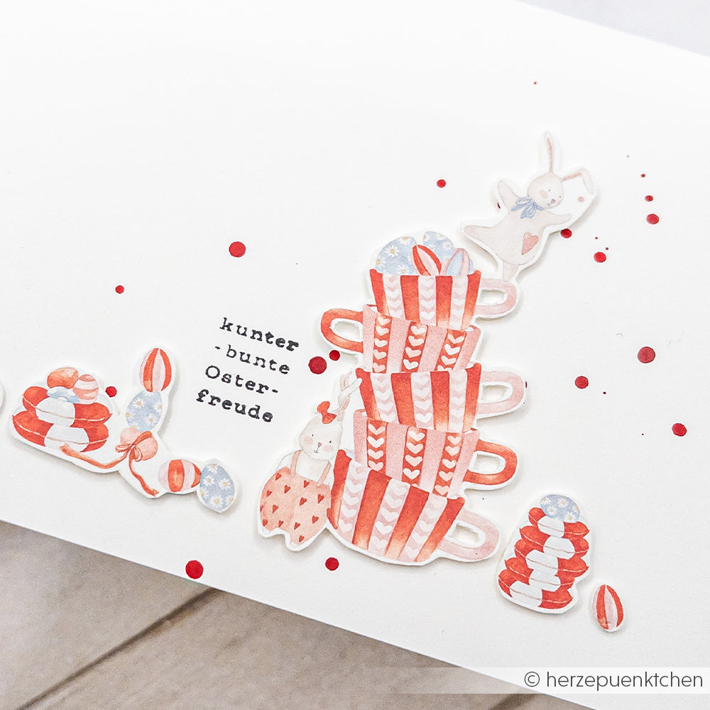 Illustration von gestapelten roten Teetassen, Hasen, Ostereiern und deutschem Text auf weißem Hintergrund - ideal für Grußkarten oder kreative Papierprojekte. Entwurf von Alexandra Renke.
