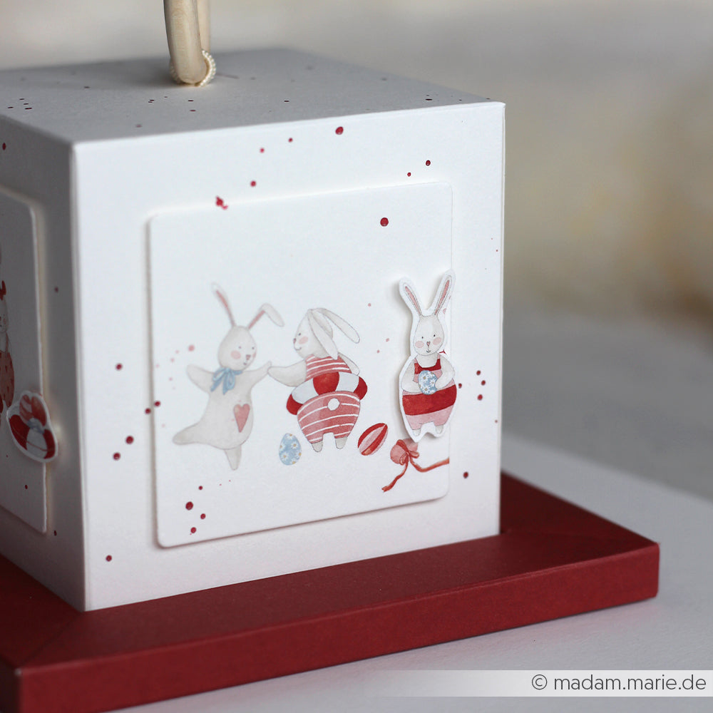 Weiße Schachtel mit rotem Boden, verziert mit verspielten Hasen, die Herzen und Ostereier in Pastellfarben halten - perfekt für Papierprojekte oder zum Ausstellen Ihres Lieblingsdesignpapiers von Alexandra Renke.