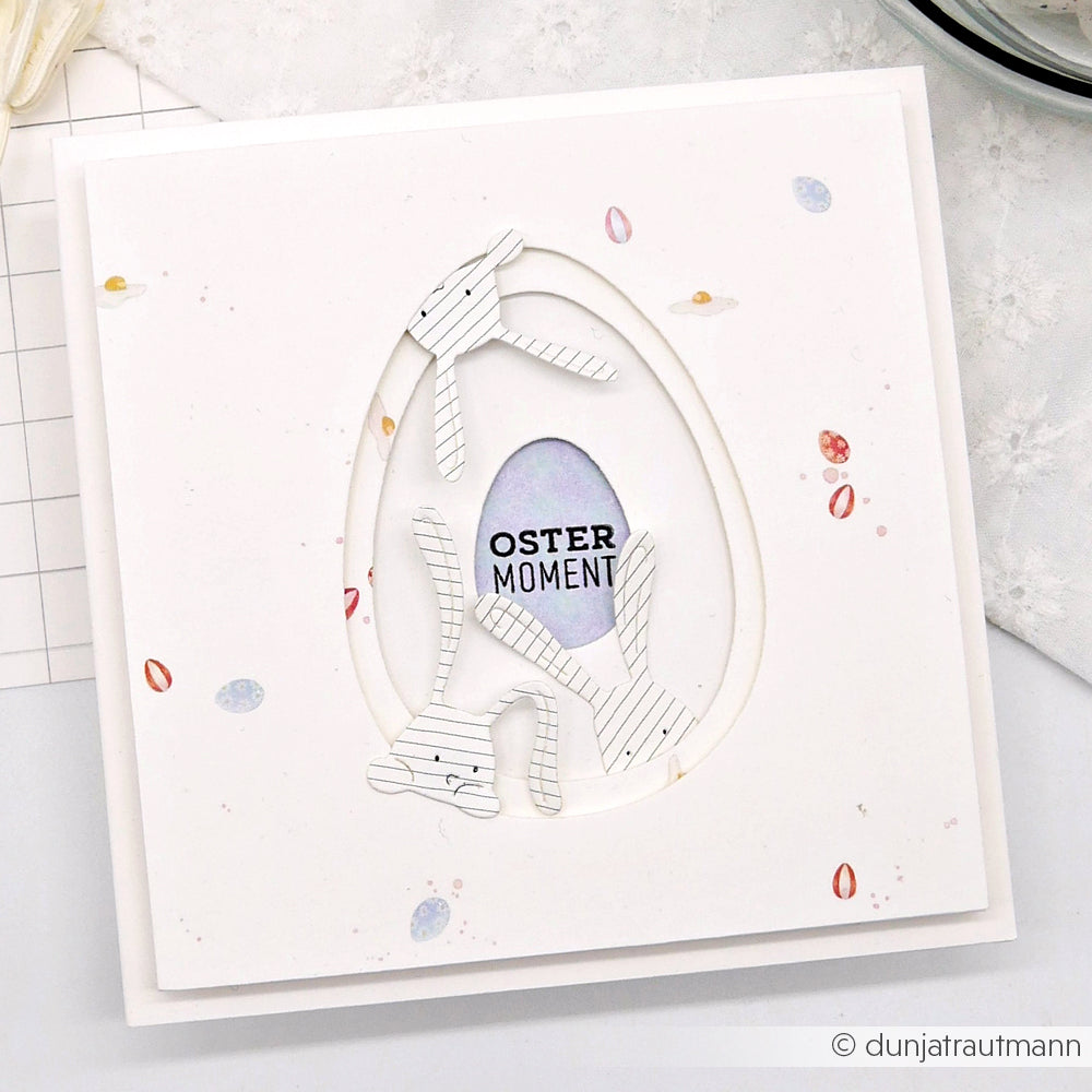 Handgefertigte Osterkarte mit dem Designpapier Ostereierreigen Bunt von Alexandra Renke, mit einem ausgeschnittenen Ei, gestreiften Hasenformen und "Oster Moment" im Inneren - eine einzigartige Papierprojekte-Kreation.