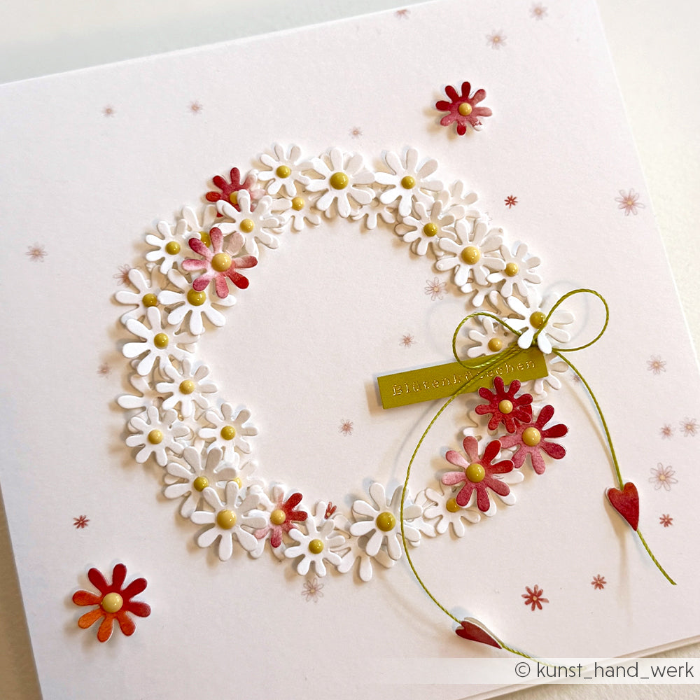 Ein Kranz aus weißen und roten Gänseblümchen schmückt eine weiße Karte mit einer Schleife und kleinen roten Herzen - ideal für Papierprojekte oder die Buchbinderei, mit dem zarten Touch des Designpapiers Blumenreigen von Alexandra Renke.