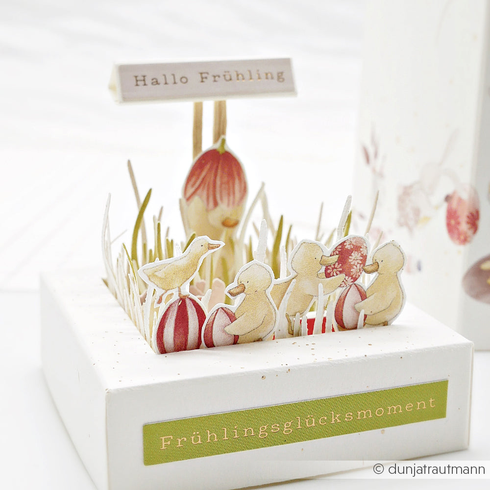 Ein frühlingshaftes Papierdiorama von Alexandra Renke mit Enten, Eiern, Gras und Schildern mit "Hallo Frühling" und "Frühlingsglücksmoment" - perfekt als Inspiration für Papierprojekte oder handgefertigte Grußkarten!.