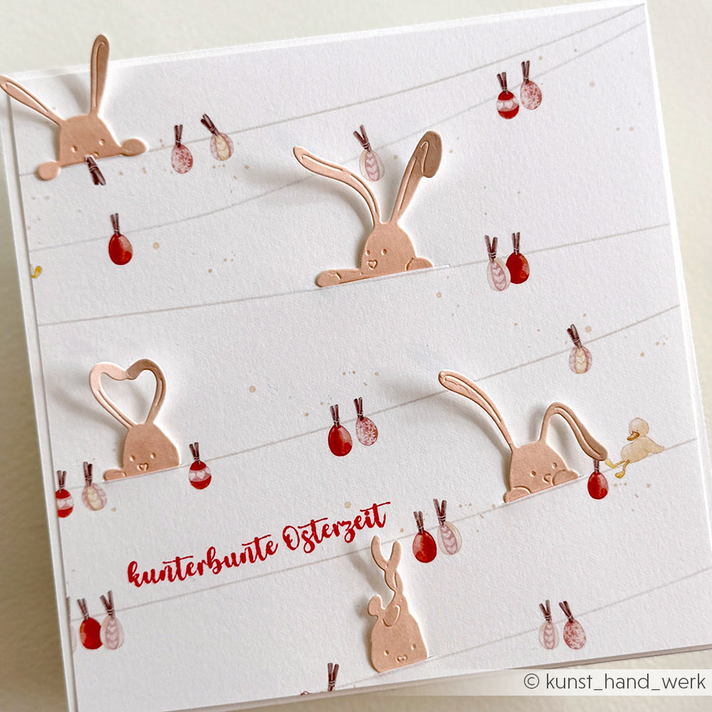 Weiße Karte von Alexandra Renke mit verspielten Kupferhasenausschnitten und kleinen gemalten Ostereiern auf der Leine, Text: kunterbunte Osterzeit - eine perfekte Inspiration für Papierprojekte.
