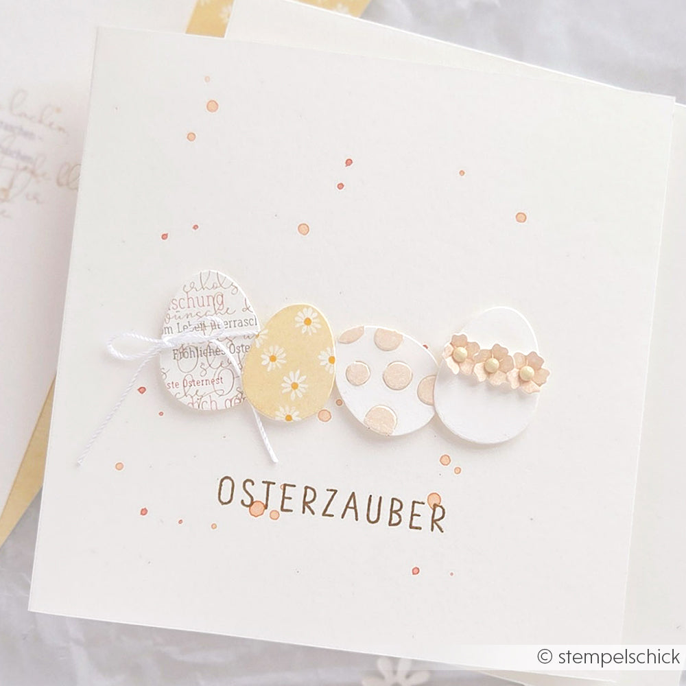 Weiße Osterkarte von Alexandra Renke mit vier verzierten Eiern, orangefarbenen Punkten und dem Text "OSTERZAUBER" - ideal für Papierprojekte oder in Verbindung mit Margeritenreigen auf Gelb Designpapier.