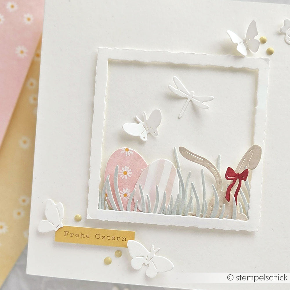 Handgemachte Osterkarte von Alexandra Renke mit pastellfarbenen Eiern, Schmetterlingen, Libelle, Gras und "Frohe Ostern"-Aufkleber. Mit charmanten Margeritenreigen auf Rosa Designpapier für einen einzigartigen Papierprojekte-Look.