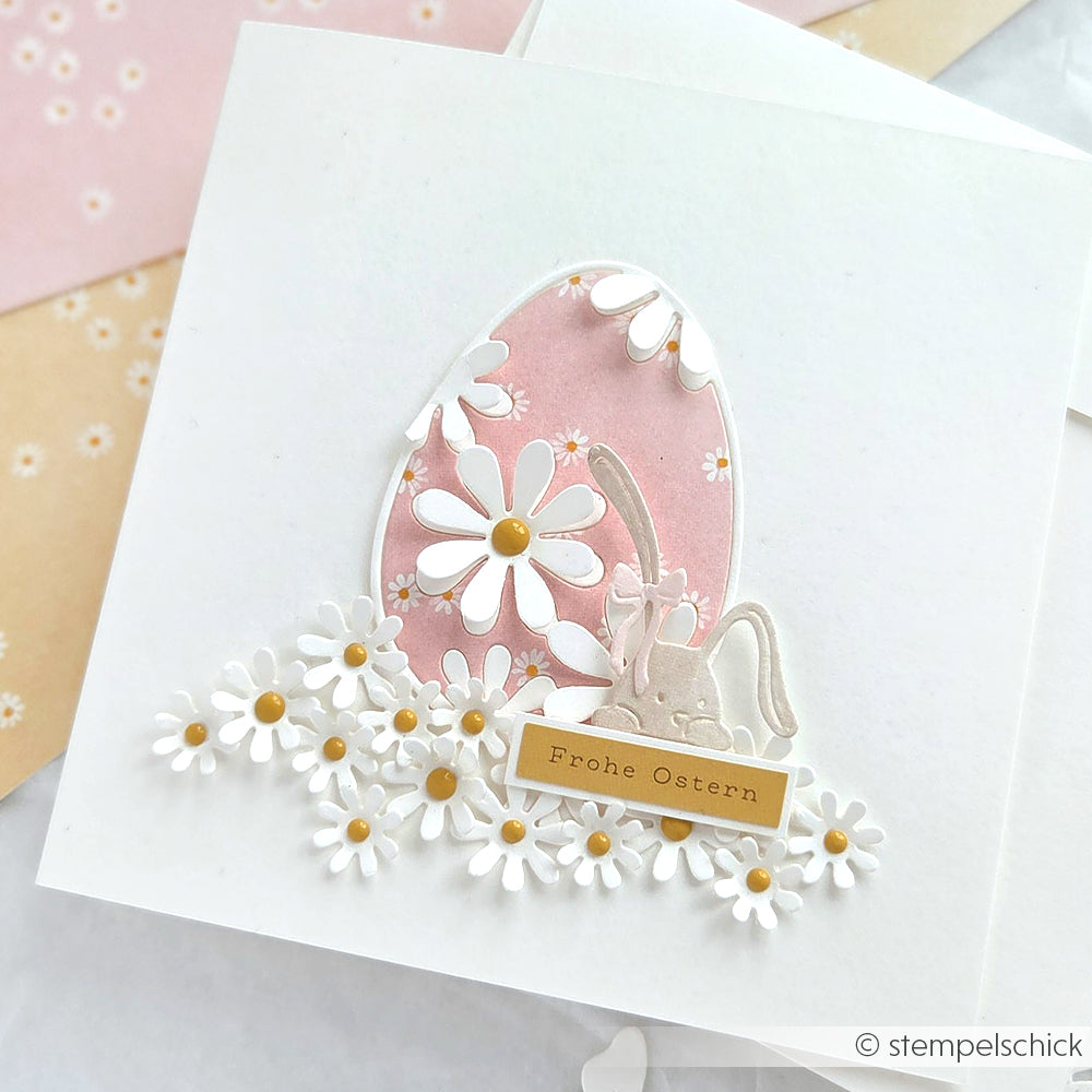 Alexandra Renke Quadratische Osterkarte mit rosa Margeritenreigen, Gänseblümchen, Hase und Ei auf Designpapier. Mit "Frohe Ostern" auf einem goldenen Etikett - ideal für festliche Grüße und kreative Papierprojekte.