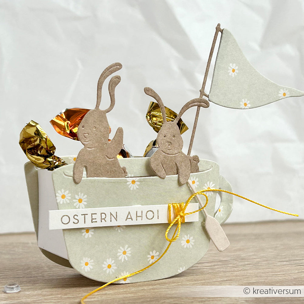 Ein Papierboot von Alexandra Renke Designpapier enthält zwei Hasenausschnitte, Pralinen, eine Flagge und einen OSTERN AHOI-Anhänger - alles wunderschön auf Holz arrangiert für entzückende Papierprojekte.