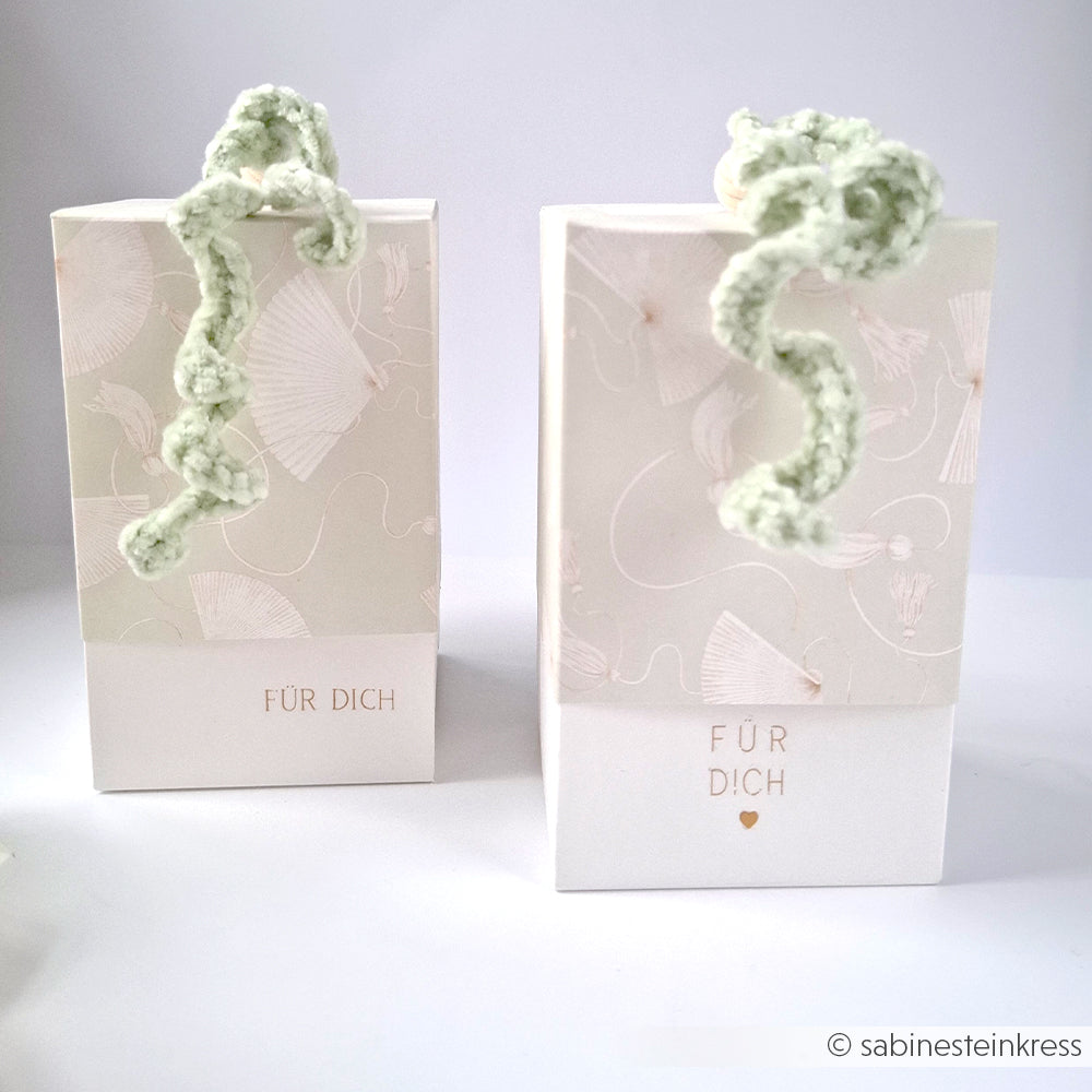 Zwei dekorative pastellfarbene Geschenkboxen von Alexandra Renke aus Designpapier mit dem Schriftzug FÜR DICH" und einem Herz auf der Vorderseite, verziert mit grünen Garnakzenten.
