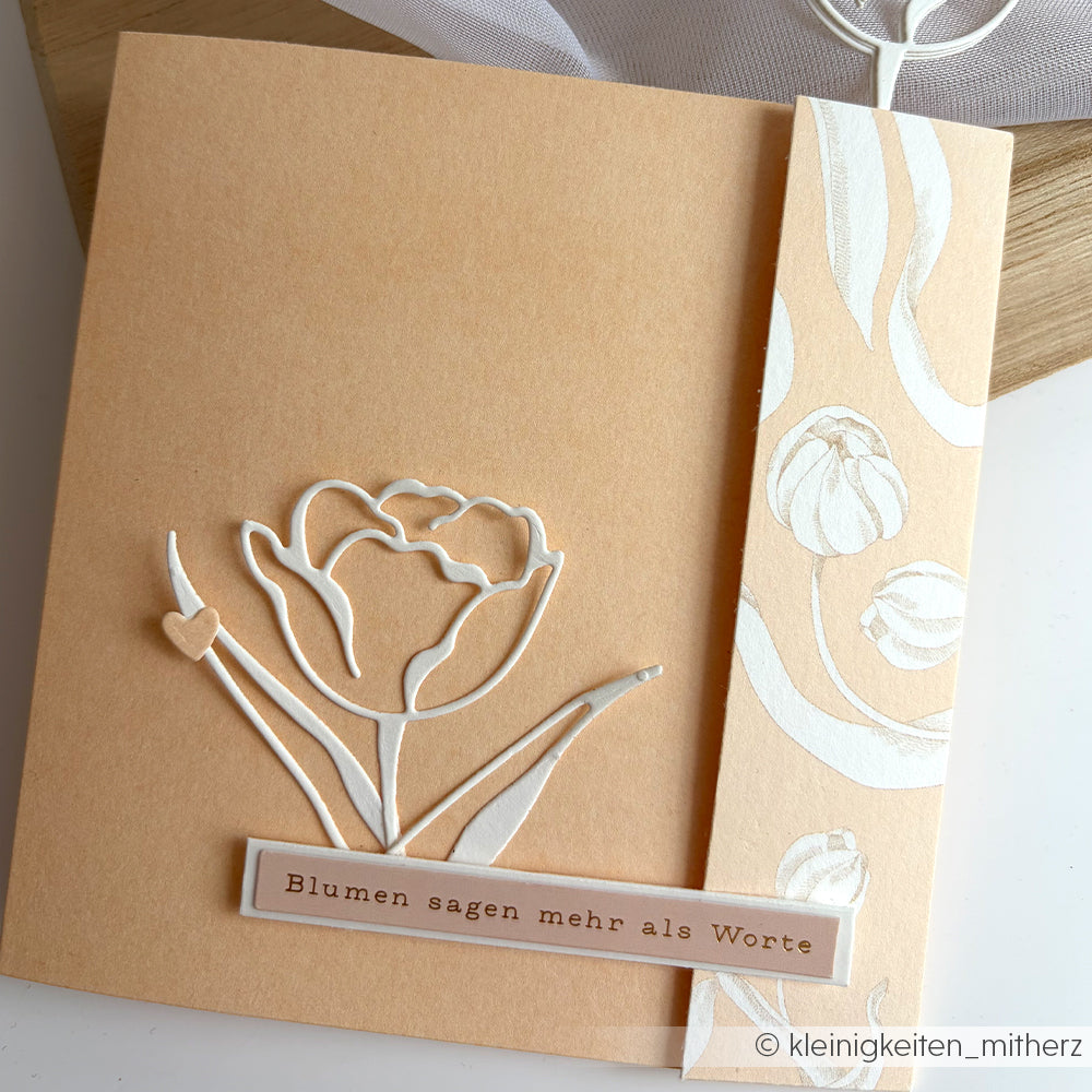 Beigefarbene Karte von Alexandra Renke mit einer weißen Blume und der Prägung "Tulpen auf Elegant-Marille Designpapier", mit der Aufschrift "Blumen sagen mehr als Worte" - perfekt für besondere Papierprojekte.
