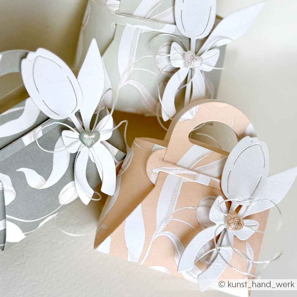 Drei dekorative Geschenkboxen von Alexandra Renke aus Designpapier mit floralen Prints und geschichteten weißen Blumen erinnern an Handtaschen - ideal für Papierprojekte oder als Geschenk für Tulpen auf Elegant-Marille.