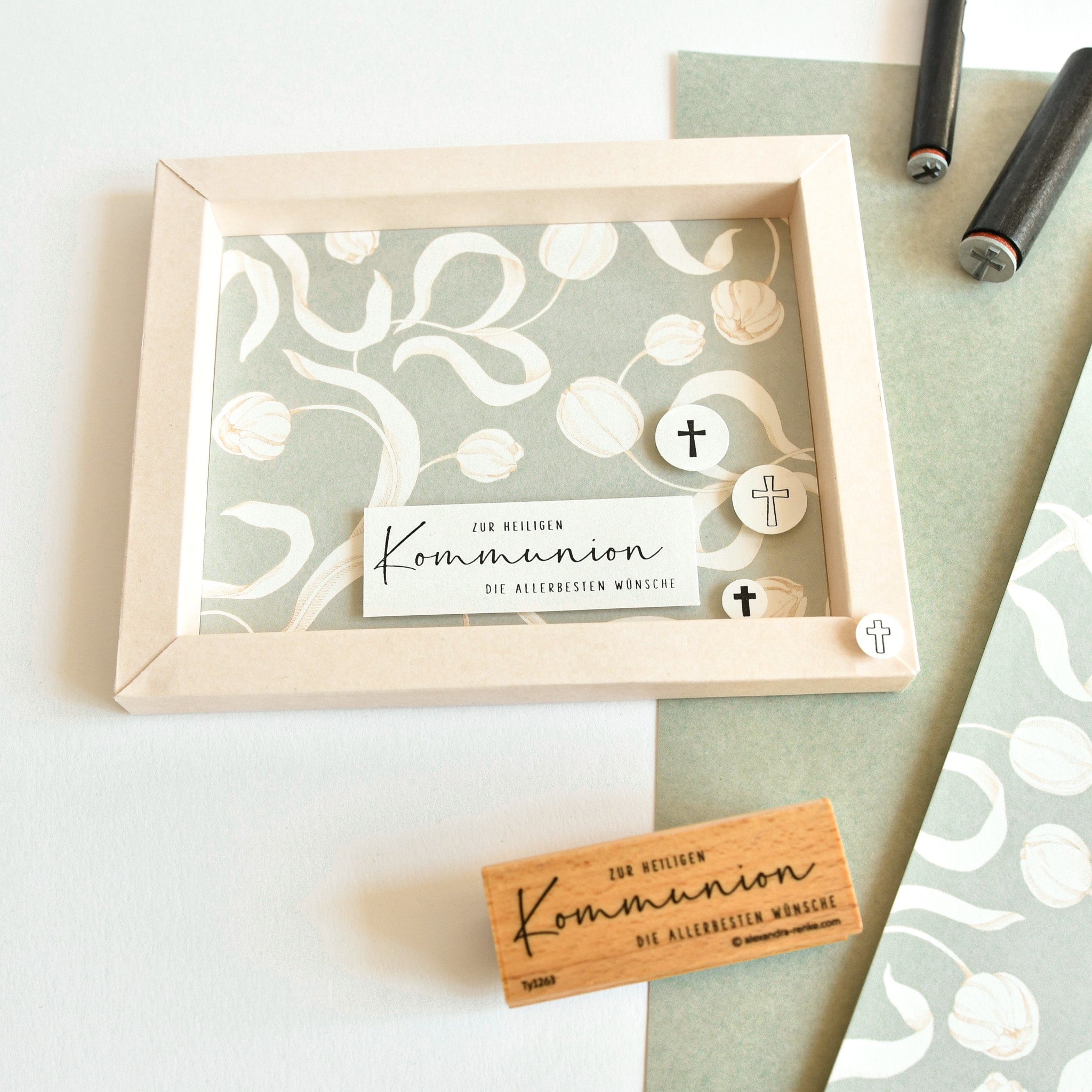 Gerahmte Karte mit Kommunionstext, Kreuzmotiven und floralem Hintergrund, hergestellt aus Alexandra Renkes Designpapier "Tulpen auf Elegant-Salvia" für distinctive Papierprojekte; abgebildet auf einem Schreibtisch mit Briefmarken und Stift.
