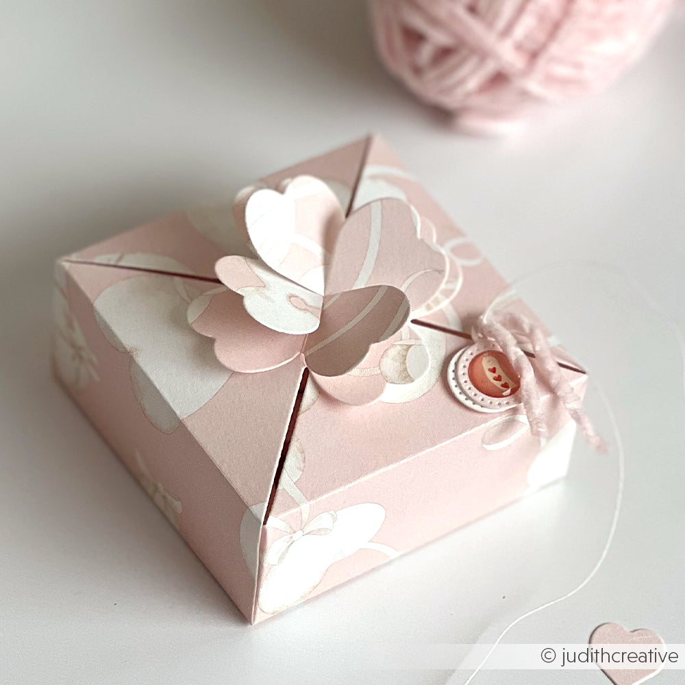 Rosa geblümte Geschenkbox mit herzförmigen Klappen aus Alexandra Renke Designpapier, Schnur und Knopf - ideal für Papierprojekte oder zum Verschenken mit einzigartigem Designpapier. Ausgestellt auf einer weißen Fläche mit rosa Garn.