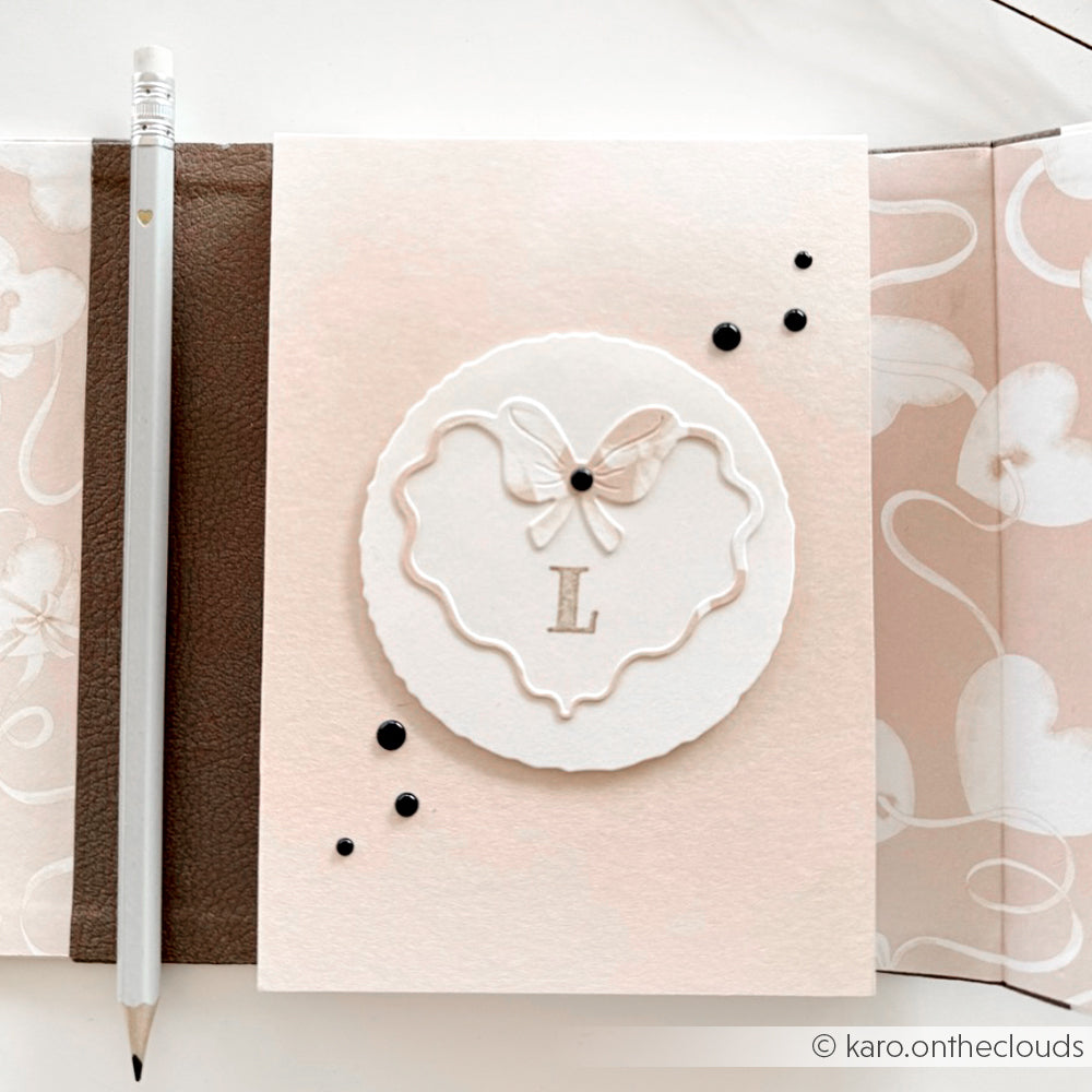 Elegante weiße Karte von Alexandra Renke mit Herzen auf Rosa, Schmetterlingsmotiv, Buchstabe L, und schwarzen Punkten - ideal für Papierprojekte oder als stilvolles Designpapier; abgebildet neben einem weißen Bleistift.