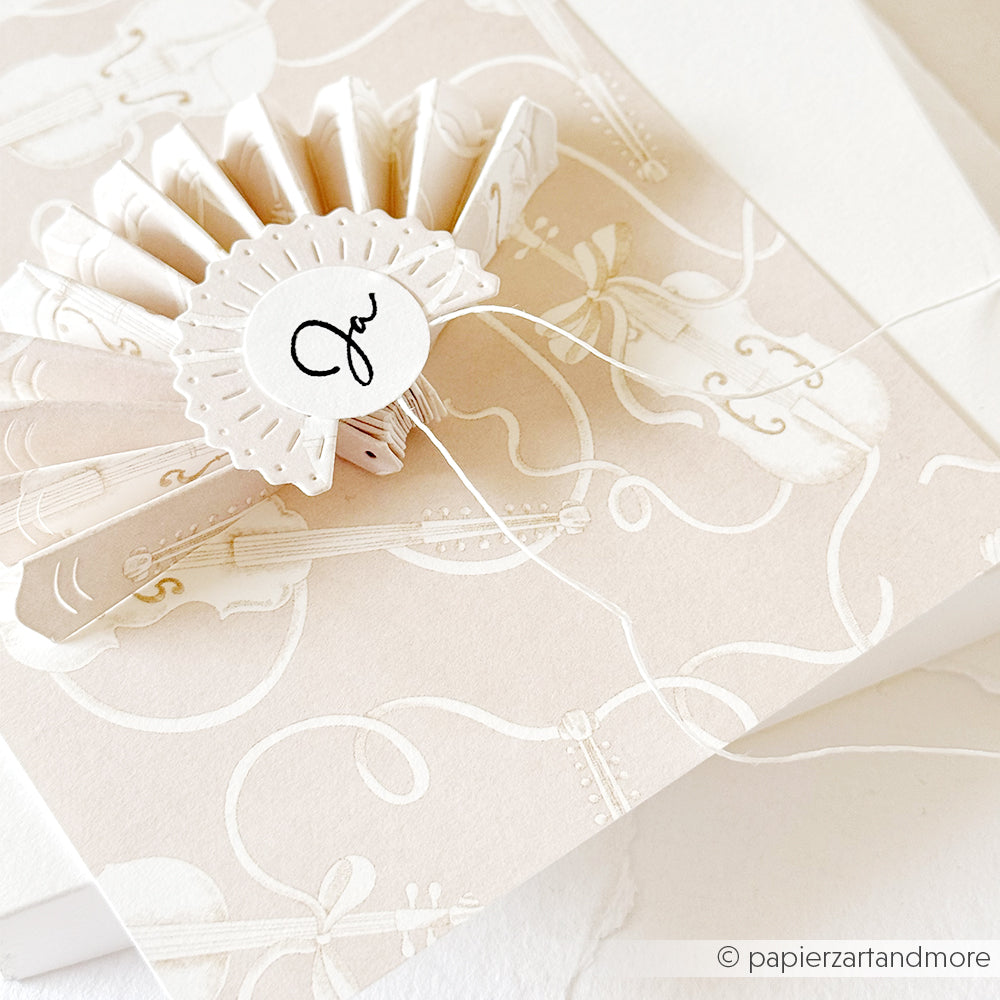 Elegante Einladung mit Rosette und "Ja" in Schreibschrift auf cremefarbenem Alexandra-Renke-Papier mit Buchbinderei-Details und Geigen-Designpapier mit Geigen- und Notenmotiven verziert.