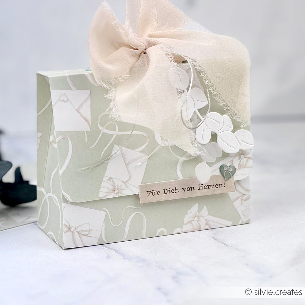 Eine pastellfarbene Geschenkbox von Alexandra Renke aus elegantem Designpapier mit einer Stoffschleife und einem "Für Dich von Herzen!"-Anhänger ruht auf einer Marmorfläche.
