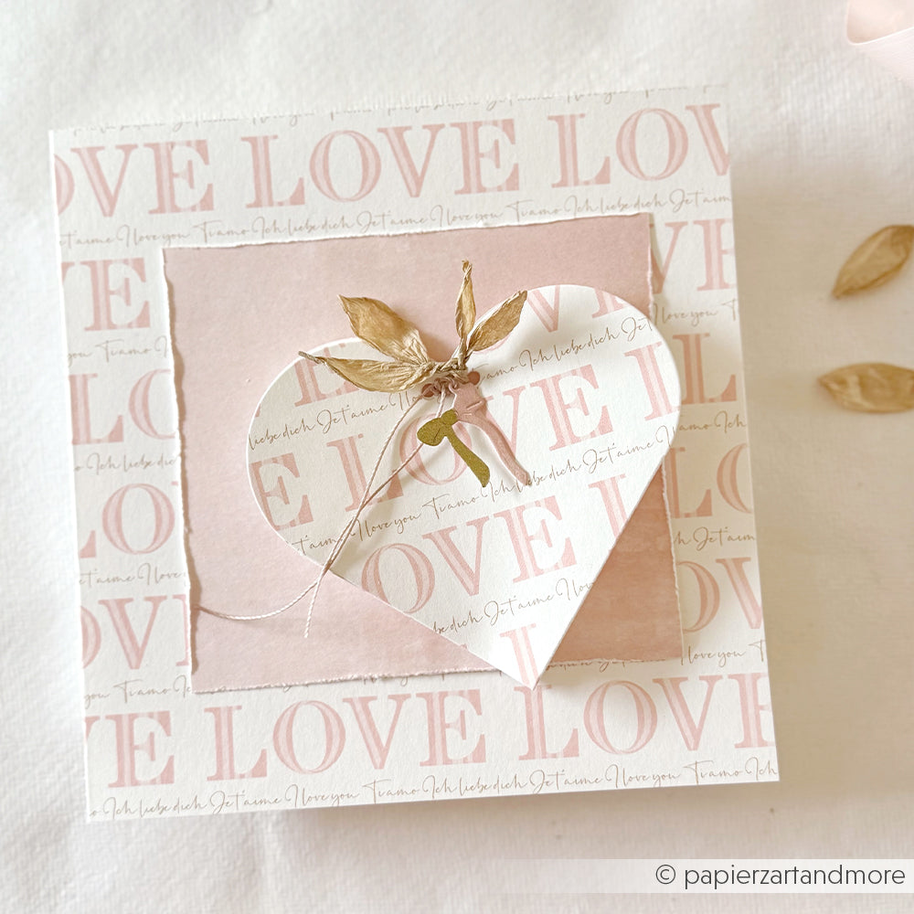 Quadratische Karte von Alexandra Renke mit dem Text "Love" in Serifentypo gestreift Rosa, einem rosa Herz, Bindfaden und getrockneten Blättern auf hellem Designpapier.