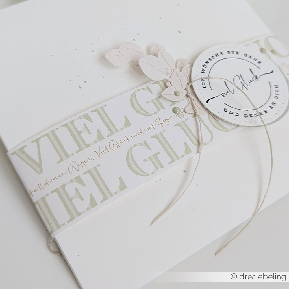 Weiße Grußkarte von Alexandra Renke mit "Viel Glück"-Banderole, beigen Blattausschnitten auf Designpapier und einem runden, handgeschriebenen Anhänger. Veredelt mit einem Hauch von Viel Glück Elegant-Grün für zusätzlichen Charme.