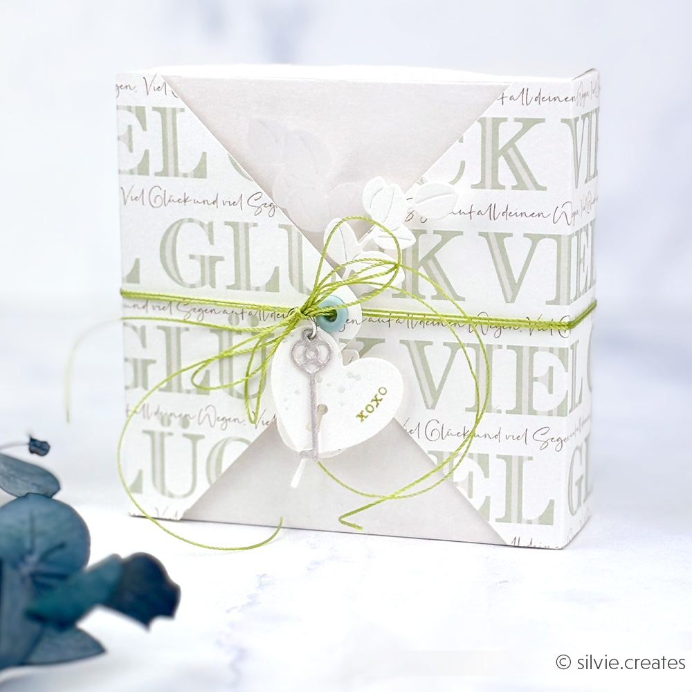 Eine weiße Geschenkbox von Alexandra Renke mit grüner Kordel, einem Herzanhänger und einem Schlüsselanhänger mit dem Motiv "GLÜCK" - ideal für Papierprojekte oder als Grußkarten-Inspiration.