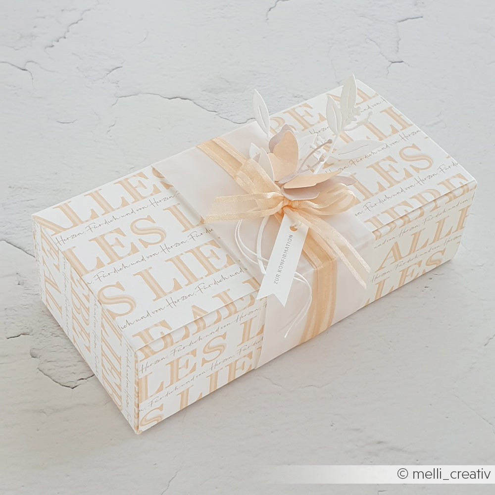 Elegante Geschenkbox von Alexandra Renke, eingewickelt in weißes und beiges Designpapier, verziert mit Schleife und dekorativen Blättern auf hellem Hintergrund.