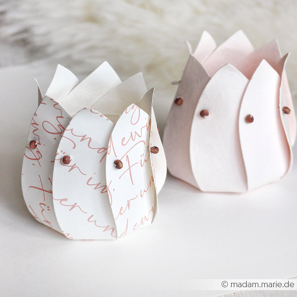 Zwei handgefertigte Lotuskerzenhalter von Alexandra Renke aus Designpapier - einer mit Schreibschrift, der andere in hellem Rosa - ruhen auf einem weichen Hintergrund. Eine ideale Inspiration für Papierprojekte oder einzigartige Grußkarten.