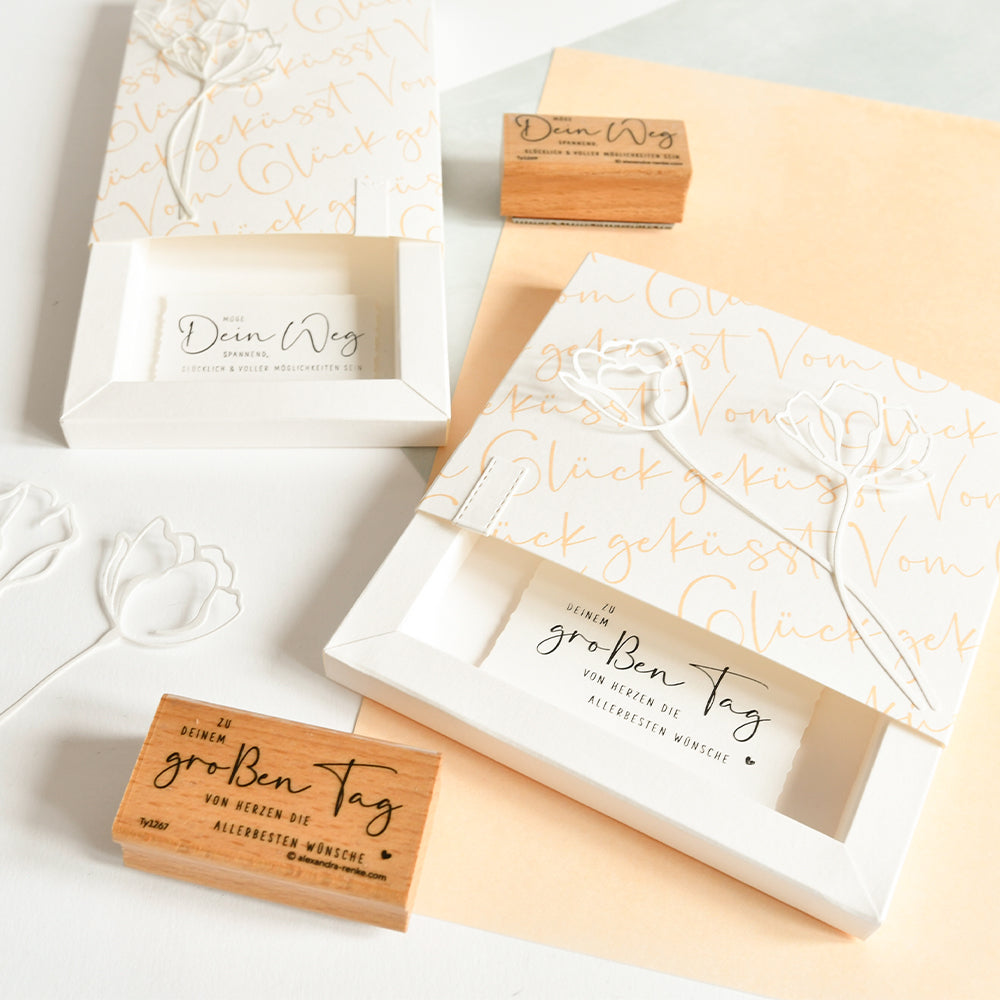 Zwei dekorative Geschenkboxen aus Alexandra Renke Designpapier mit floralen Motiven, dazu zwei kleine Typo Vom Glück geküsst Elegant-Marille Holzstempel, auf hellem Untergrund - ideal für Papierprojekte.