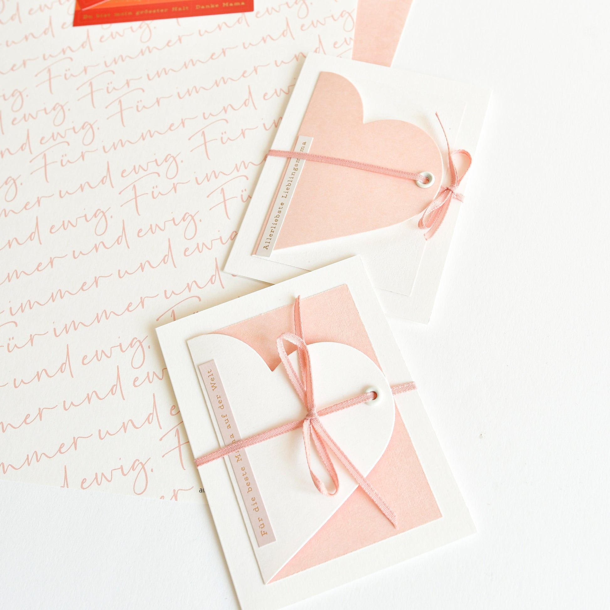 Zwei rosafarbene und weiße, mit rosafarbenem Band gebundene Karten in Herzform, hergestellt mit Alexandra Renke Elegant-Rosa Designpapier, werden auf einem weißen Hintergrund mit handgeschriebenem Text dargestellt - ideal für einzigartige Papierprojekte.