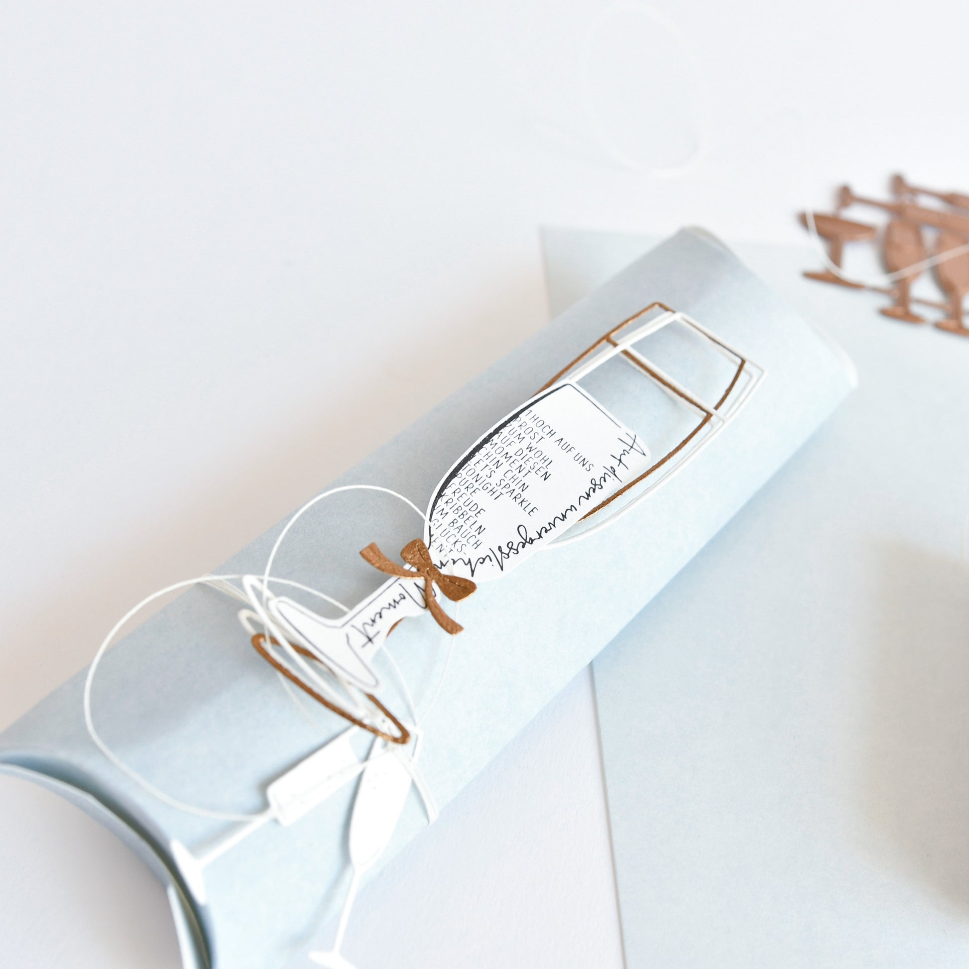 Hellblaues Alexandra Renke Mimi Elegant-Blau hell Designpapier, gerollt und mit weißer Kordel gebunden, mit dekorativem Sektglasausschnitt und eleganter Schrift - ideal für einzigartige Papierprojekte.