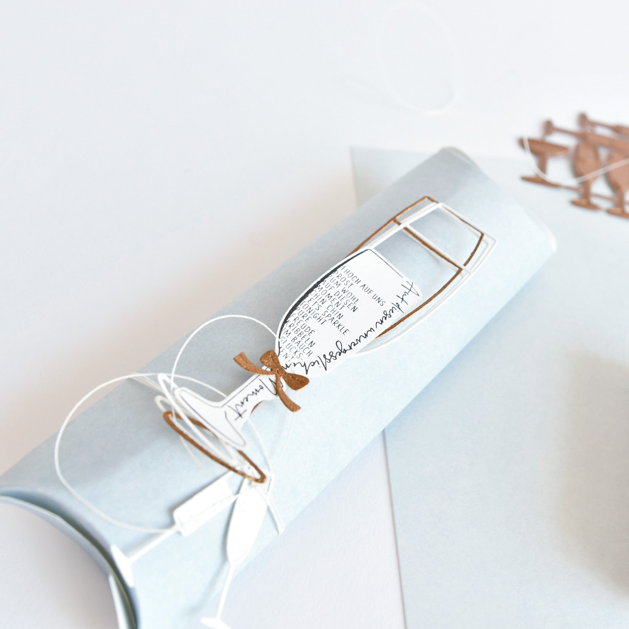 Hellblaues Alexandra Renke Mimi Elegant-Blau hell Designpapier, gerollt und mit weißer Kordel gebunden, mit dekorativem Sektglasausschnitt und eleganter Schrift - ideal für einzigartige Papierprojekte.