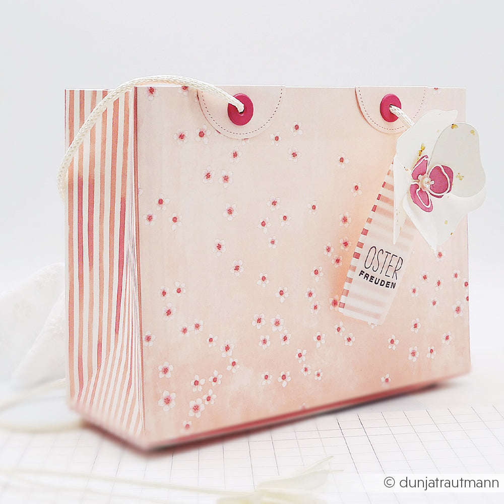 Rosa-weiß geblümte Geschenktasche mit gestreiften Seiten, Schmetterlingsanhänger und Kordelgriffen - perfekt für Alexandra Renke Papierprojekte mit Kirschblüten Designpapier auf hellem Hintergrund.